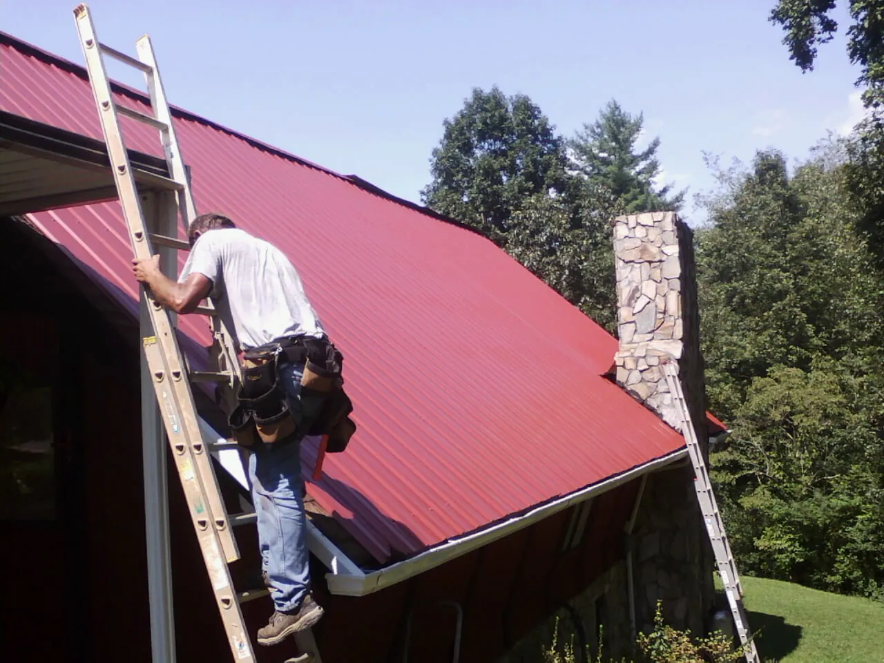 Roofer on a ladder beside a red metal roof and stone chimney — Soffit & Fascia Repair by Roofing Brothers in Paisley